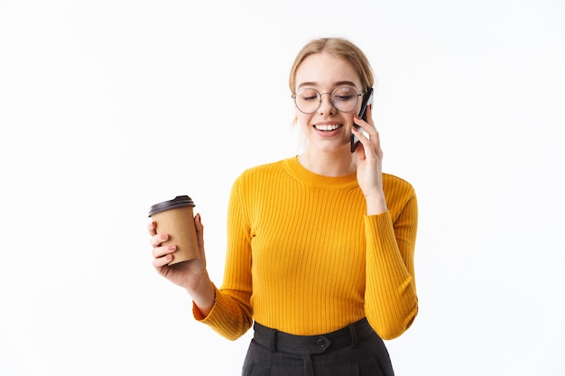 Attraente giovane donna bionda che indossa un maglione in piedi isolato su un muro bianco, parlando al telefono cellulare, tenendo una tazza di caffè da asporto