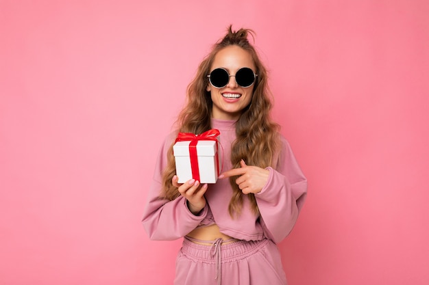 Attraente giovane donna bionda riccia sorridente felice isolata su una parete di fondo rosa che indossa abiti sportivi rosa e occhiali da sole che tengono scatola regalo guardando la fotocamera