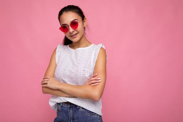 Attraente giovane donna bruna che indossa abiti eleganti di tutti i giorni e occhiali da sole moderni isolati su una parete di fondo colorata guardando la telecamera