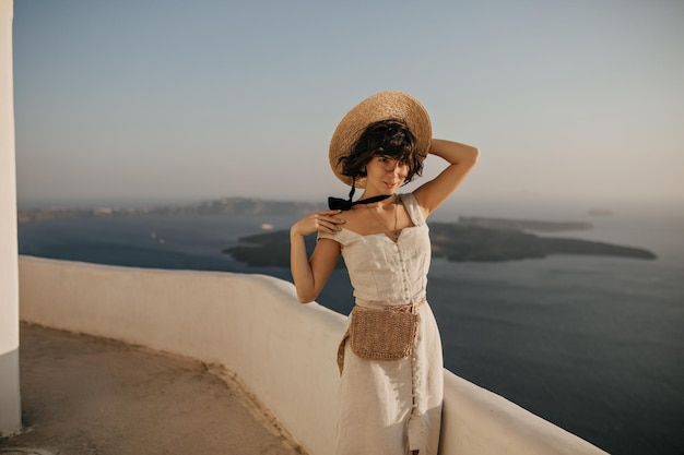 Attraente giovane donna bruna in abito beige e cappello di paglia posa in un posto bellissimo con vista mare Ritratto di signora riccia in abito elegante all'esterno