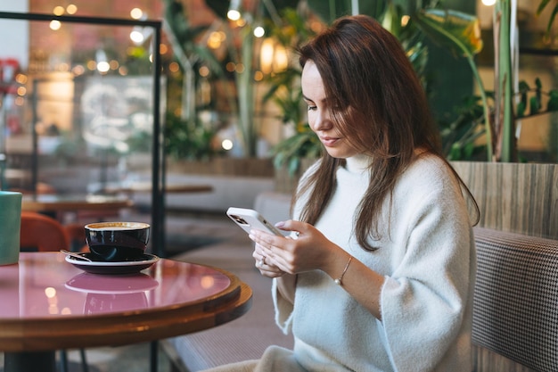 Attraente giovane donna bruna in abito casual bianco con una tazza di caffè utilizzando il telefono cellulare nella caffetteria