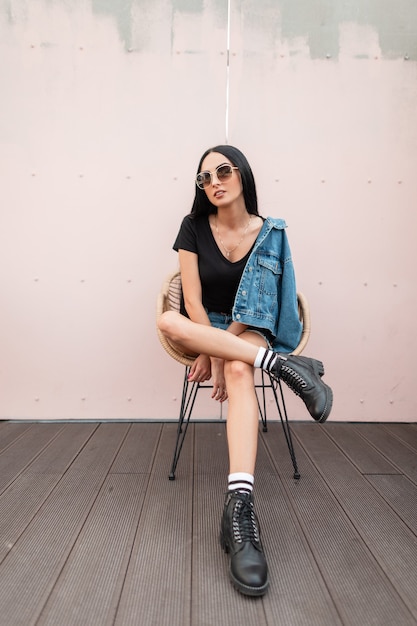 Attraente giovane donna bruna in una maglietta in una giacca di jeans in occhiali da sole in stivali in pelle nera si siede su una sedia vicino al muro sulla strada in città. Ragazza alla moda hipster all'aperto. Stile autunnale