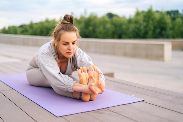 Attraente giovane donna che fa esercizio di pratica yoga in posa seduta piegata in avanti seduta su tappetino da yoga nel parco cittadino Yogini in forma sportiva femminile che indossa abbigliamento sportivo facendo esercizi di stretching all'esterno