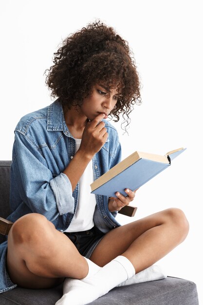 Attraente giovane donna che indossa denim seduto su una sedia isolata sul muro bianco, leggendo un libro di carta