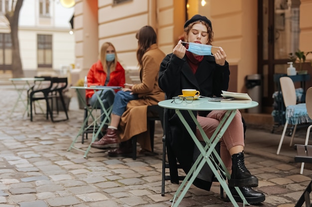 Attraente giovane donna che toglie maschera medica per bere caffè caldo al bar locale.