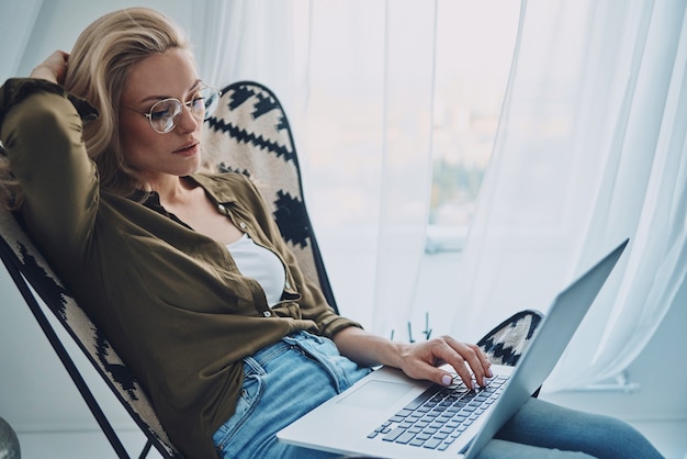 Attraente giovane donna che utilizza il computer portatile mentre è seduto su una sedia comoda a casa