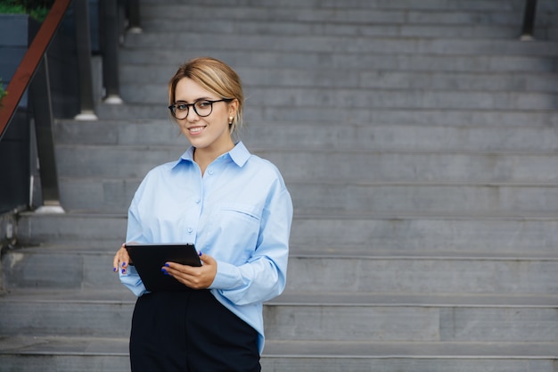 Attraente giovane donna con i capelli biondi utilizzando tablet digitale mentre in piedi sulle scale vicino edificio per uffici. Persone di concetto, affari e tecnologia moderna.