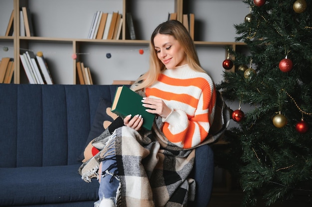 Attraente giovane donna con i capelli scuri che legge un libro interessante mentre si siede sul divano grigio Sfocatura dello sfondo del bellissimo albero di natale Atmosfera accogliente