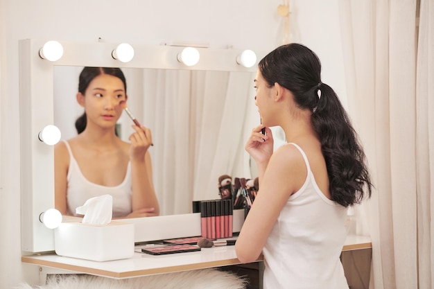 Attraente giovane donna con il trucco realizzato da un artista professionista che si guarda allo specchio al chiuso