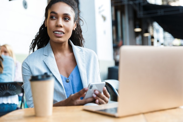 Attraente giovane donna d'affari africana sorridente seduta al tavolo del bar all'aperto, lavorando su un computer portatile