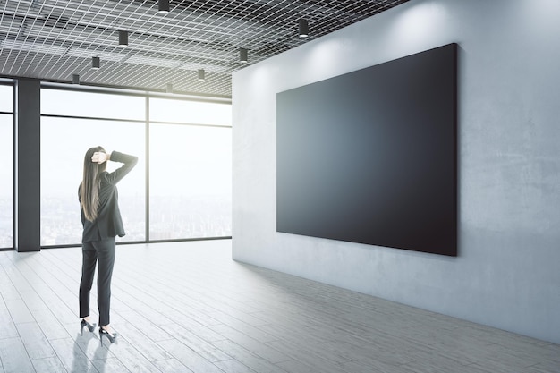 Attraente giovane donna d'affari europea riflessiva che guarda uno striscione nero vuoto all'interno con vista sulla città e luce diurna