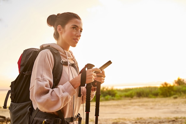 Attraente giovane donna escursionista in felpa con smartphone e bastoncini da trekking