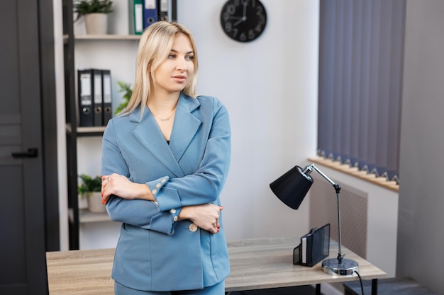 Attraente giovane donna in abito blu in piedi vicino al tavolo mentre guarda la fotocamera Giovane donna professionale in posa sul posto di lavoro in ufficio