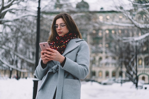 Attraente giovane donna in abito invernale sfogliando smartphone in strada con la neve