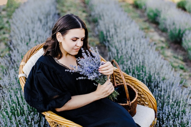 Attraente giovane donna in abito nero seduto in poltrona circondato da campo di lavanda e tenendo cesto con fiori