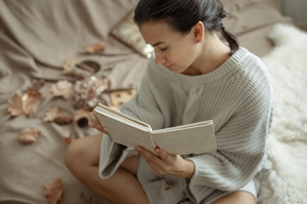 Attraente giovane donna in accogliente maglione lavorato a maglia leggendo un libro a letto.