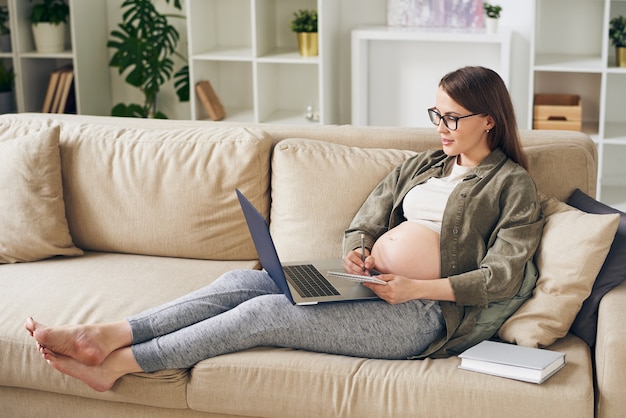 Attraente giovane donna incinta in homewear seduto sul divano e leggere l'articolo sul laptop mentre prende appunti sulla maternità