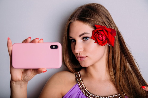 Attraente giovane donna prende un selfie sul suo smartphone, imbronciato le labbra e raddrizzandosi i capelli con un fiore di rosa rossa.