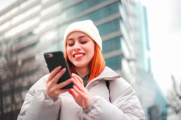 Attraente giovane donna rossa che digita un messaggio tramite il suo telefono cellulare in una città che sta sorridendo