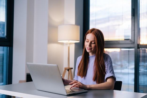 Attraente giovane donna rossa che lavora con il computer portatile al tavolo in un ufficio moderno seduto alla scrivania sullo sfondo della finestra con il paesaggio urbano.