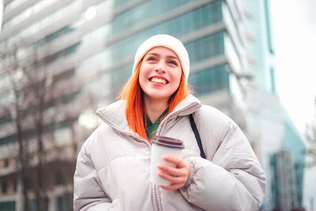 Attraente giovane donna rossa sorridente e guardando al cielo un caffè da asporto in mano