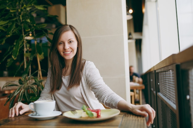 Attraente giovane donna seduta da sola vicino alla grande finestra nella caffetteria al tavolo con una tazza di cappuccino, torta, rilassante nel ristorante durante il tempo libero. Giovane femmina che ha resto in caffè. Concetto di stile di vita.