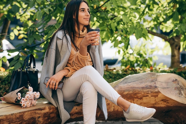 Attraente giovane donna seduta sulla panchina e bere caffè nel parco Donna nella strada della città che beve cappuccino caldo Bella ragazza bruna con una tazza di caffè Caffè in movimento