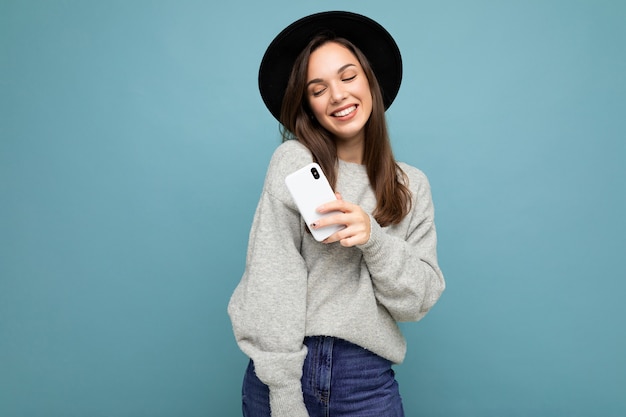 Attraente giovane donna sorridente che indossa cappello nero e maglione grigio tenendo lo smartphone guardando verso il basso isolato su sfondo.spazio copia