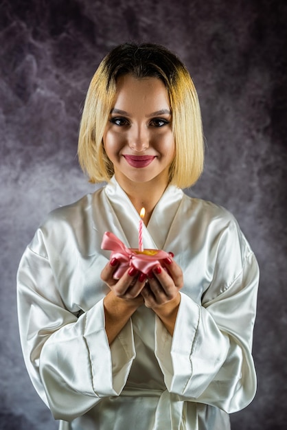 Attraente giovane donna sorridente eccitata con il trucco e in accappatoio che tiene il bigné di compleanno con la candela isolata su un semplice sfondo compleanno in un accappatoio