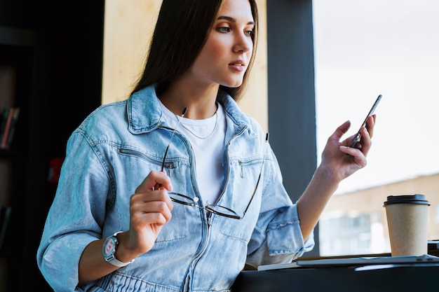 Attraente giovane donna sta vicino alla finestra, tiene lo smartphone in mano, occhiali per la visione e lavora sul computer portatile.