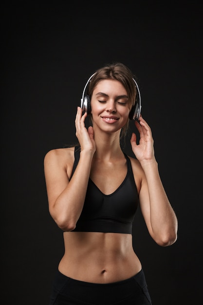 Attraente giovane e sana fitness donna che indossa reggiseno sportivo e pantaloncini isolati su sfondo nero, ascoltando musica con le cuffie