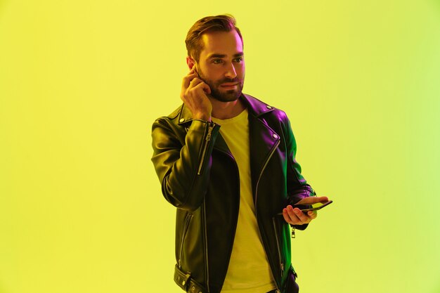 Attraente giovane fiducioso che indossa una giacca di pelle in piedi isolato su un muro giallo, ascoltando musica con auricolari wireless mentre tiene il cellulare