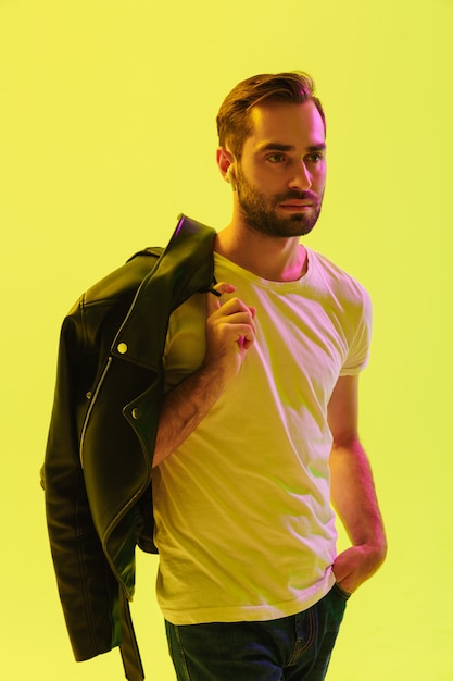 Attraente giovane in piedi isolato su un muro giallo, con in mano una giacca di pelle sulla spalla