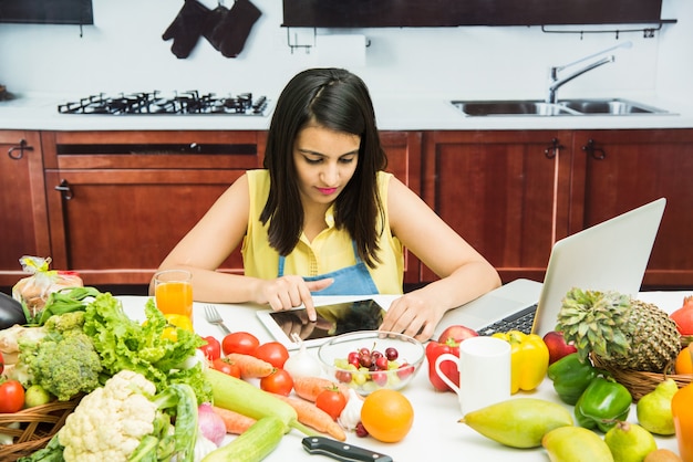 Attraente giovane ragazza indiana o donna con grembiule in cucina multitasking, utilizzando smartphone, tablet pc con tavolo pieno di frutta e verdura e computer