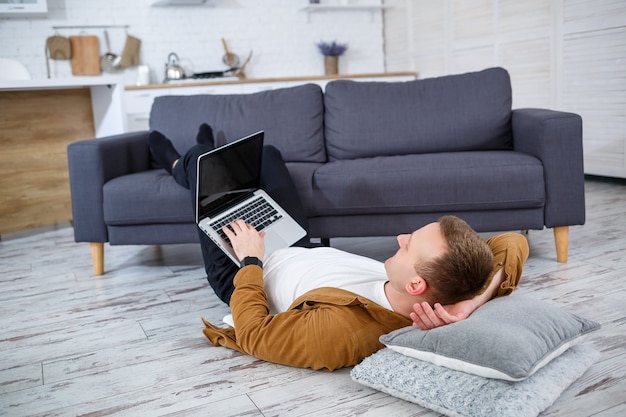 Attraente giovane sorridente in abbigliamento casual seduto sul pavimento sul divano in soggiorno utilizzando il computer portatile