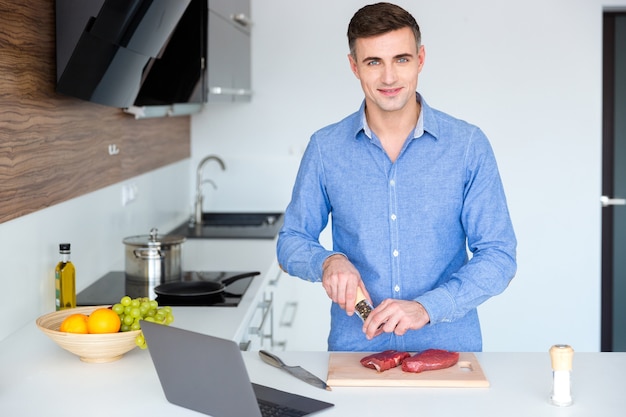 Attraente giovane sorridente in merda blu che cucina carne in cucina a casa