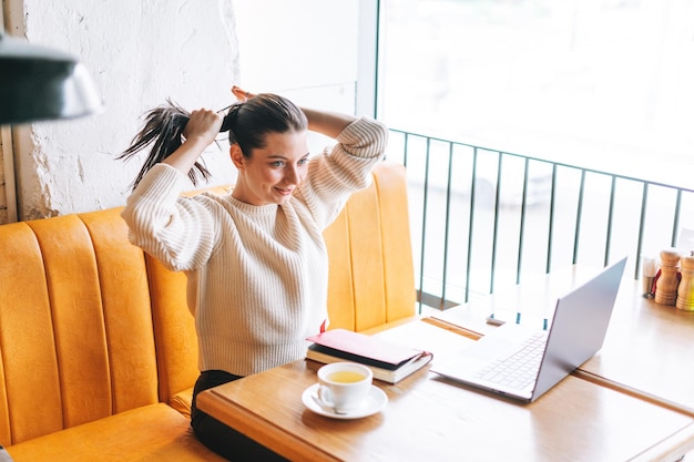 Attraente giovane studentessa sorridente bruna freelance con una tazza di tè utilizzando il computer portatile nella caffetteria