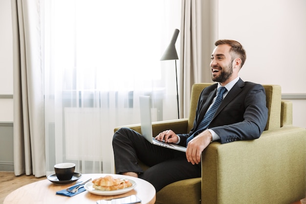 Attraente giovane uomo d'affari che indossa un abito seduto su una sedia nella camera d'albergo, lavorando su un computer portatile mentre fa colazione