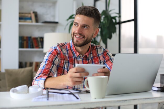 Attraente giovane uomo seduto alla scrivania in ufficio a casa e utilizzando il telefono cellulare per controllare le reti sociali.