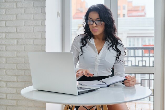 Attraente imprenditrice lavorando con computer portatile, seduto alla scrivania vicino alla finestra. Donna alla moda in camicia bianca che guarda il monitor