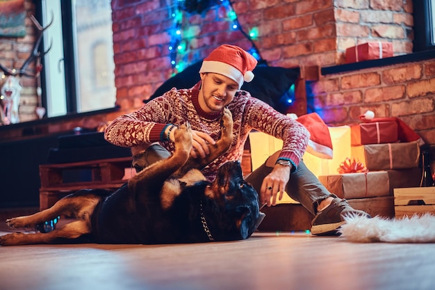 Attraente maschio hipster barbuto si siede su un pavimento con il suo cane Rottweiler in una stanza con decorazioni natalizie.