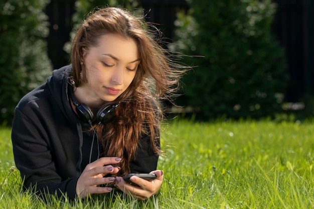 Attraente modella bruna con i capelli lunghi vestita in felpa con cappuccio nera sdraiato sul prato verde guardando smartphone
