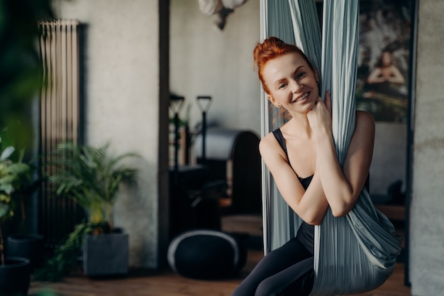 Attraente piccola donna dai capelli rossi con un bel sorriso seduto in posizione comoda su un'amaca appesa al soffitto nel centro fitness e in attesa di una lezione di yoga antigravità. Stile di vita sano e sport