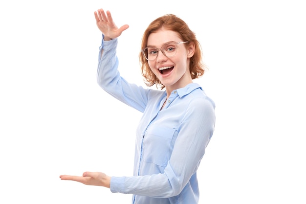 Attraente promotore della ragazza rossa in camicia blu da lavoro che tiene qualcosa in mano dimostrando lo spazio vuoto con la faccia eccitata isolata su sfondo bianco