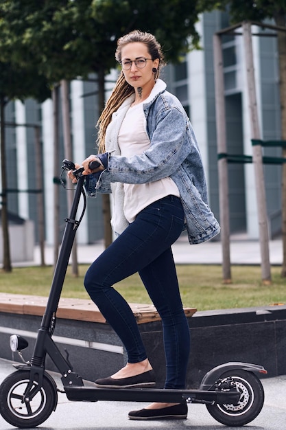 Attraente ragazza alla moda con i dreadlocks sta guidando uno scooter elettrico per strada vicino a un edificio interessante.