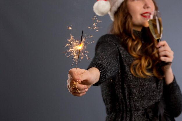 Attraente ragazza allegra con i capelli ricci in cappello di Natale sorridente azienda sparkler e bere champagne isolato su sfondo grigio