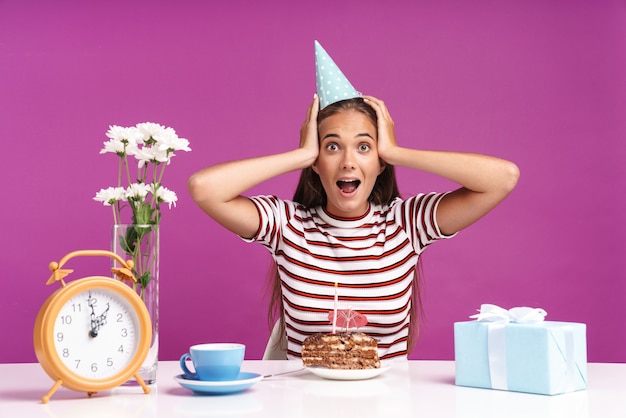 Attraente ragazza allegra seduta alla scrivania con una torta di compleanno isolata sul muro rosa, festeggiando