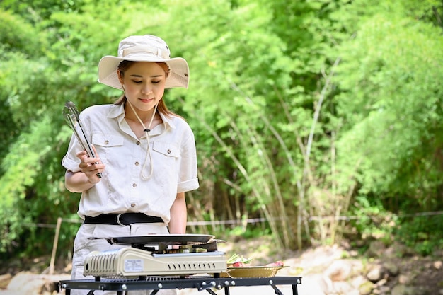 Attraente ragazza asiatica che prepara un cibo da picnic in campeggio utilizzando un fornello da picnic per grigliare una bistecca di manzo