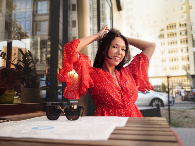 Attraente ragazza asiatica con un vestito rosso si appunta i capelli sulla terrazza estiva di un caffè.