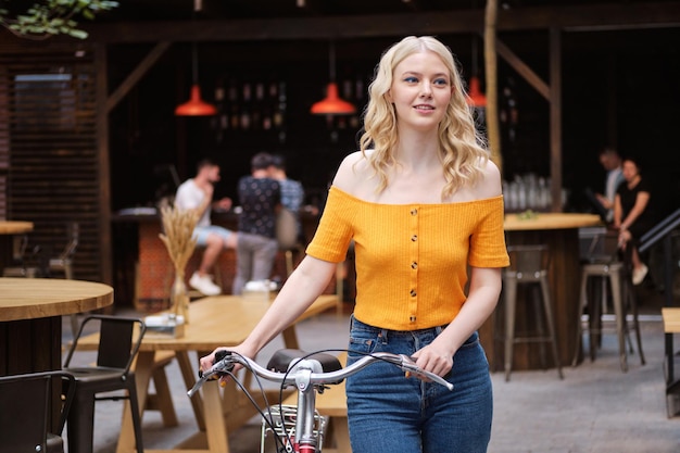 Attraente ragazza bionda casual in piedi con la bicicletta classica nel cortile del caffè della città